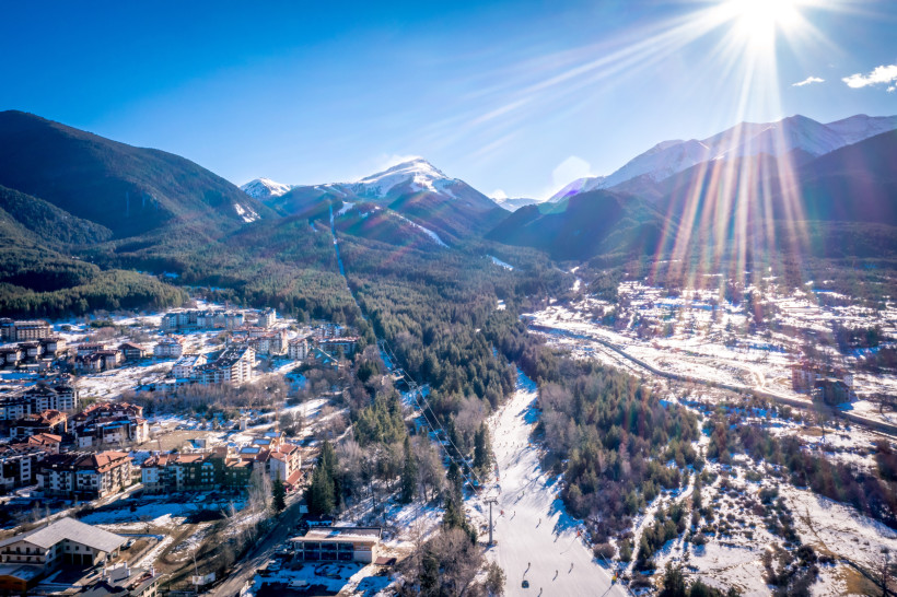Winterliche Luftaufnahme von Bansko mit Blick auf eine belebte Skipiste, die sich durch dichte Wälder zieht. Umgeben von verschneiten Bergen des Pirin-Gebirges, leuchtet die tiefstehende Sonne mit langen Lichtstrahlen über der Szene. Am unteren Bildrand s