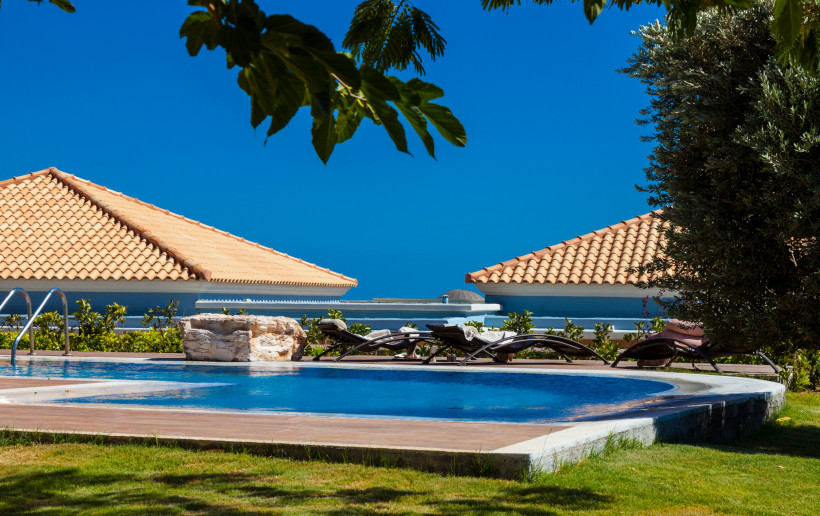 Das Bild zeigt einen gepflegten Poolbereich in einer modernen Hotelanlage.    Der runde Pool ist von einer Holzterrasse umgeben, auf der mehrere elegante Liegestühle mit weißen Handtüchern bereitstehen.    Im Hintergrund sind zwei Gebäude mit typischen Te