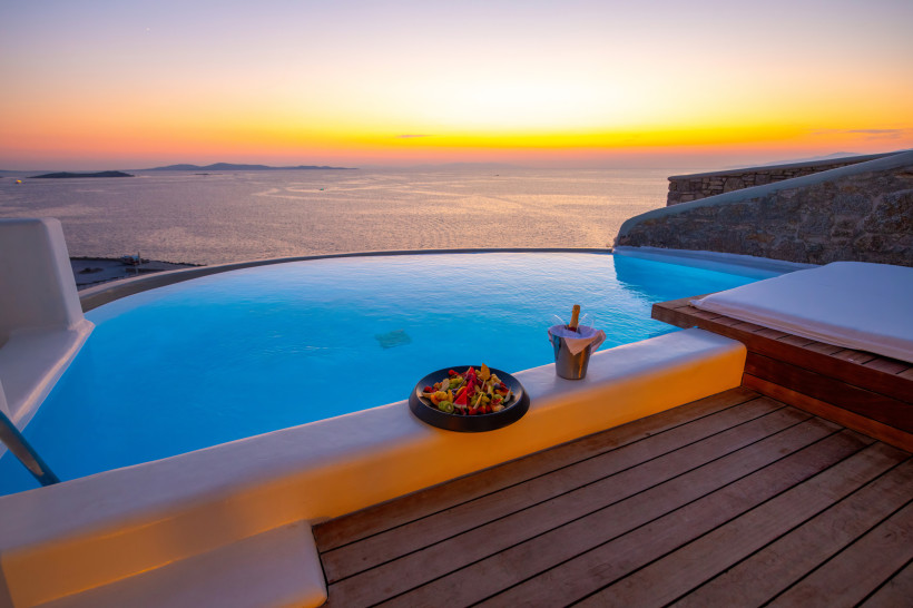 Infinity-Pool auf einer Terrasse mit Blick auf das ruhige Meer bei Sonnenuntergang. Auf dem Rand des Pools stehen ein Teller mit frischem Obst und ein Eiskübel mit einer Flasche Sekt. Der Himmel leuchtet in warmen Gelb- und Orangetönen.