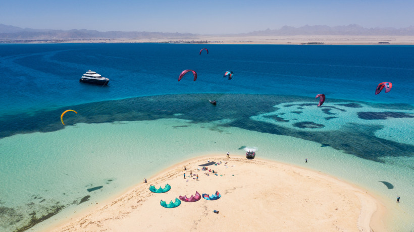 Ägypten Bunte Kites schweben über türkisfarbenem Wasser vor einer Sandbank im Roten Meer. Auf der Sandbank liegen mehrere Kites und Menschen bereiten sich auf den Wassersport vor. Im Hintergrund ankert ein Boot, dahinter liegt das Festland mit einer kargen Küsten