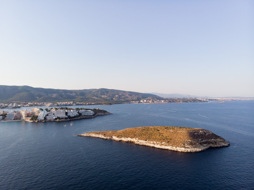 Blick auf die Isla de sa Porrassa vor der Küste Mallorcas mit Bucht und Küstenbebauung im Hintergrund