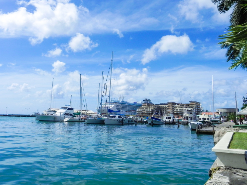 Blick auf den Jachthafen von Oranjestad, Aruba. Mehrere Motorboote und Segelyachten liegen im ruhigen Wasser vertäut. Im Hintergrund steht ein großes Kreuzfahrtschiff vor modernen Gebäuden. Palmen und blauer Himmel runden die Szene ab.