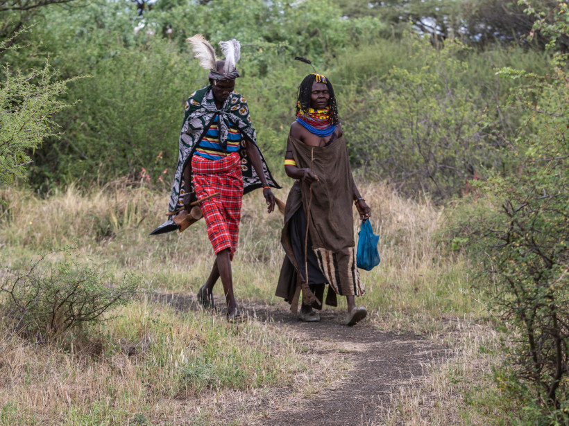Kenia Zwei Angehörige der Samburu-Volksgruppe gehen auf einem schmalen Pfad durch eine bewachsene Savannenlandschaft. Der Mann trägt eine rote karierte Wickelhose, farbige Kleidung und Federschmuck am Kopf, die Frau trägt eine traditionelle braune Robe mit bunt