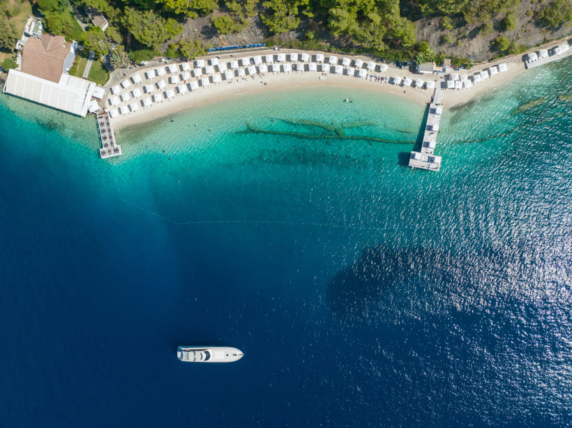 Luftaufnahme eines luxuriösen Strandabschnitts mit weißen Cabanas entlang eines hellen Sandstrands. Zwei Stege führen ins türkisfarbene Wasser, eine Yacht liegt weiter draußen im tiefblauen Meer vor Anker. Im Hintergrund dichter Pinienwald.