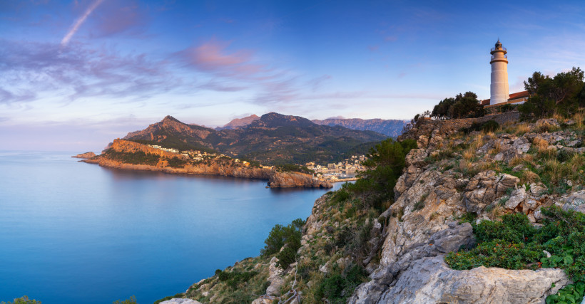 Leuchtturm Far del Cap Gros auf felsiger Küste mit Blick über eine Bucht und das Gebirge im Hintergrund