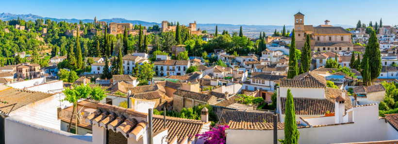 Blick über die Dächer von Granada mit der maurischen Festungsanlage Alhambra auf einem Hügel, umgeben von grüner Vegetation.
