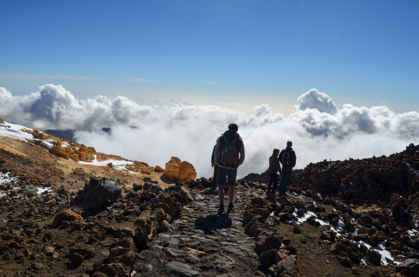 Wanderer am Teide – Über den Wolken auf Teneriffa Eine Gruppe von Wanderern steht auf einem steinigen Bergpfad am Vulkan Teide und blickt über eine dichte Wolkendecke, die wie ein weißer Teppich unter ihnen liegt. Umgeben sind sie von dunklem Lavagestein, einzelnen Schneeresten und einer kargen Hochgebir
