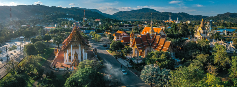 Thailand - Phuket Luftaufnahme einer eindrucksvollen Tempelanlage auf Phuket mit mehreren goldverzierten Gebäuden, roten Dächern und üppigem Grün, eingerahmt von Hügeln und blauem Himmel.