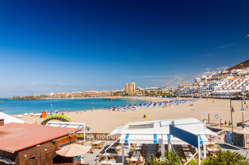 Blick auf den beliebten Strand Playa de las Vistas in Los Cristianos Panoramablick auf den Playa de las Vistas in Los Cristianos auf Teneriffa: Im Vordergrund befinden sich Terrassen und Strandrestaurants, dahinter der breite Sandstrand mit zahlreichen blauen Sonnenliegen und Sonnenschirmen. Am Ufer baden Urlauber im ruhig