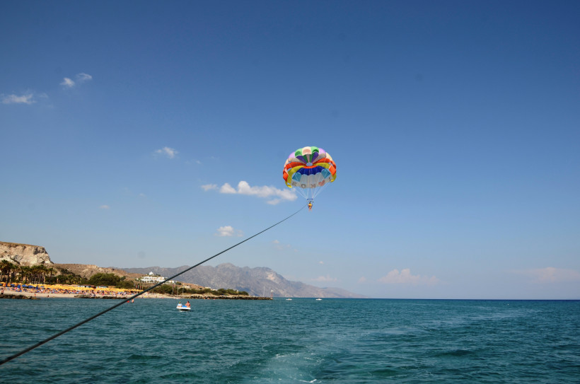 Eine Person schwebt beim Parasailing an einem bunten Fallschirm über dem türkisblauen Meer. Der Fallschirm ist über ein Seil mit einem Boot verbunden, das außerhalb des Bildes liegt. Am linken Bildrand ist ein belebter Sandstrand mit Sonnenschirmen zu seh