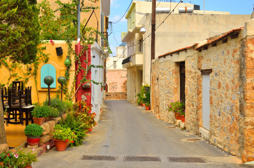 Schmale Gasse im Old Village Malia mit Taverna, Natursteinfassaden, Pflanzen und bunten Fensterläden