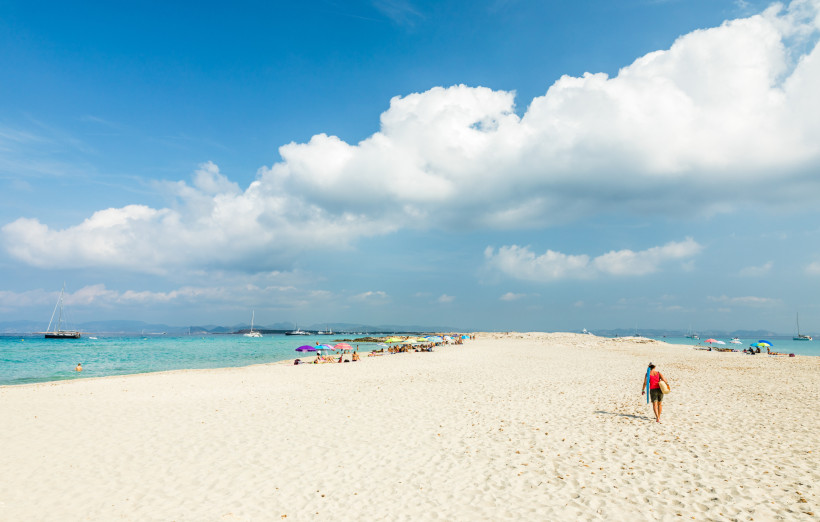 Formentera Weißer Sandstrand mit wenigen bunten Sonnenschirmen und Badenden. Eine Person geht mit einem Badetuch über der Schulter in Richtung Wasser.