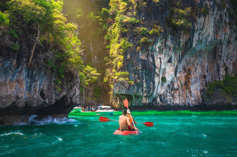 Thailand - Phuket Zwei Personen paddeln in einem roten Kajak durch türkisfarbenes Wasser in einer felsigen Bucht auf Phuket. Umgeben von hohen, mit tropischen Pflanzen bewachsenen Kalksteinfelsen fällt Sonnenlicht dramatisch durch das Blattwerk. Im Hintergrund liegen mehre