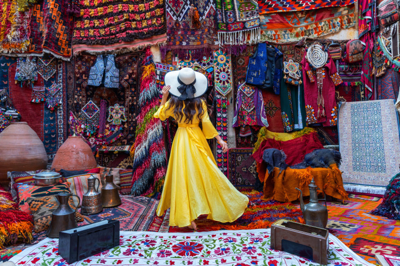 Eine Frau in einem langen, gelben Kleid und mit einem weißen Sonnenhut steht inmitten eines traditionellen orientalischen Markts mit bunten Teppichen, Stoffen, Kleidungsstücken und Kupferwaren. Sie betrachtet die kunstvoll dekorierten Textilien an der Wan