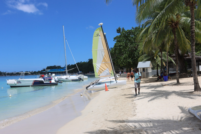 Jamaika Strand in Jamaika mit Segelbooten und Palmen. Am weißen Sandstrand liegen mehrere Boote im türkisblauen Wasser, darunter ein Katamaran mit farbigem Segel. Eine Person spaziert barfuß am Ufer entlang, während andere unter Palmen im Schatten entspannen. Im