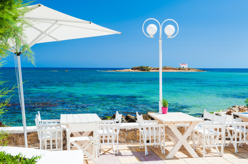 Terrasse mit weißen Tischen und Stühlen am Meer bei Malia Beach, Kapellen-Insel im Hintergrund