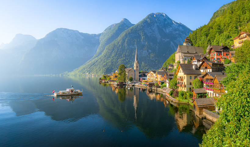 Österreich Malerisches Alpenpanorama mit dem österreichischen Ort Hallstatt am Ufer eines ruhigen Sees. Links fährt ein Ausflugsboot mit österreichischer Flagge über das spiegelglatte Wasser. Rechts am Ufer reihen sich bunte Häuser mit Holzbalkonen und Blumenschmuck