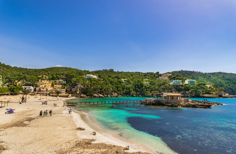 Bucht mit Sandstrand und türkisfarbenem Wasser an der Platja de Camp de Mar, mit Steg zu einem Gebäude auf einer Felsinsel