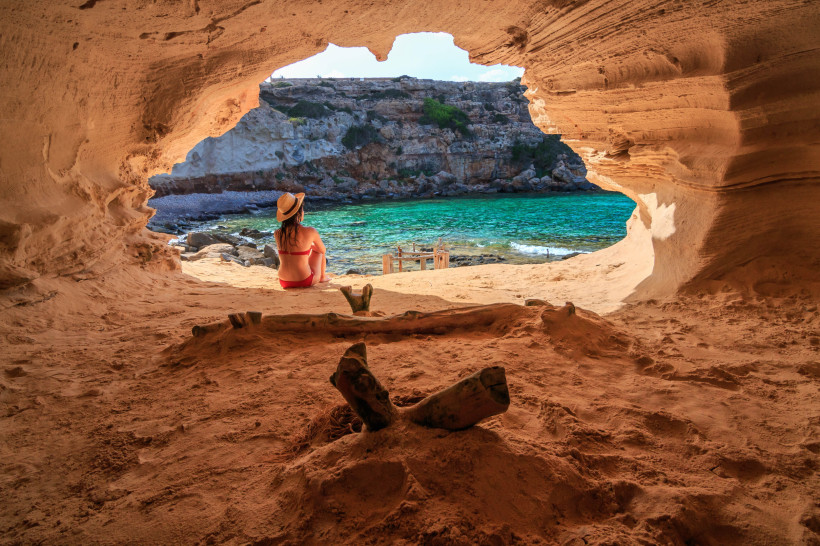 Formentera Eine Frau in einem roten Bikini und Strohhut sitzt in einer natürlichen Sandsteinhöhle und blickt aufs türkisfarbene Meer hinaus. Die Höhle öffnet sich zu einer kleinen, geschützten Bucht mit Felsen und einem Holzsteg. Die Szene vermittelt Ruhe und Naturv