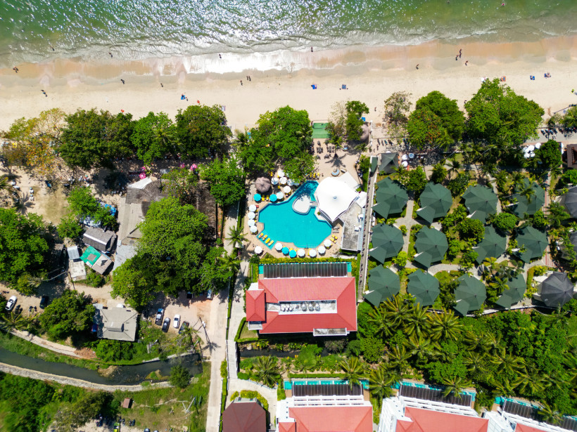 Thailand - Krabi Das Bild zeigt eine senkrechte Luftaufnahme eines tropischen Strandresorts in Thailand. Im Zentrum befindet sich ein großer, organisch geformter Swimmingpool mit hellblauen Liegen und weißen Sonnenschirmen ringsherum. Direkt dahinter erstreckt sich ein he