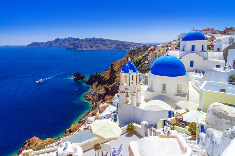 Weiße Gebäude und Kirche mit blauen Kuppeln an der Steilküste auf Santorin mit Blick auf das Meer