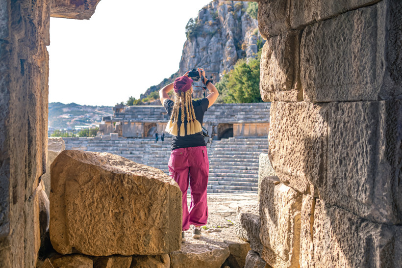 Eine Person mit langen, geflochtenen Haaren fotografiert ein antikes Amphitheater. Sie steht in einem steinernen Torbogen, im Hintergrund sind Stufen und Felsen zu sehen