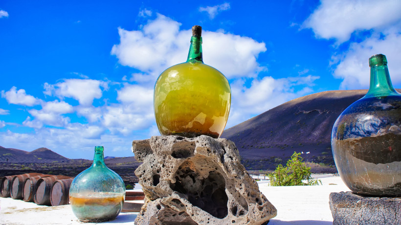Lanzarote Große grüne Glasflasche, die auf einem Lava-Steinblock steht, vor einem blauen Himmel und vulkanischen Hügeln im Hintergrund. Weitere Flaschen und Holzfässer sind sichtbar.