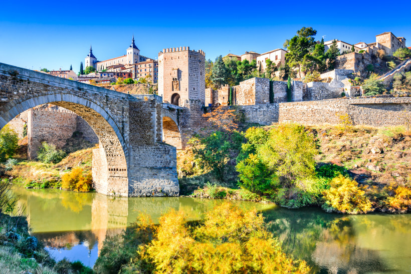 Steinerne Brücke Puente de San Martín über den Fluss Tajo in Toledo, Spanien. Im Hintergrund ist die mittelalterliche Altstadt mit dem Alcázar von Toledo zu sehen. Herbstliche Bäume spiegeln sich im ruhigen Wasser.