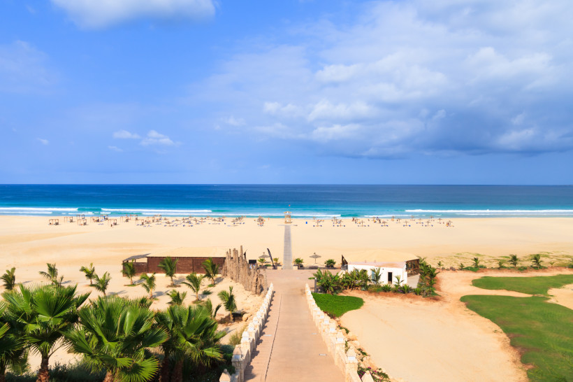 Breiter, heller Sandstrand auf der kapverdischen Insel Boa Vista mit Blick auf das tiefblaue Meer. Sonnenliegen und Schirme sind über den Strand verteilt, ein gerader Weg führt von einem Hotelgelände mit Palmen durch den Sand bis ans Wasser. Der Himmel is