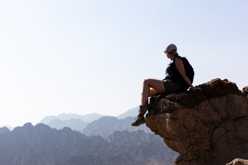 Fujairah, Aktivferien Wanderin mit Rucksack sitzt auf einem Felsvorsprung und blickt auf eine Berglandschaft