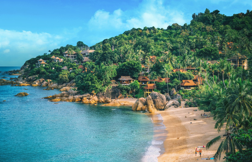 Thailand - Koh Samui Das Bild zeigt eine malerische Bucht auf Koh Samui mit einem goldfarbenen Sandstrand, türkisblauem Meer und einer von Palmen gesäumten Küste. Am Hang über dem Strand liegen zahlreiche traditionelle Villen und Bungalows mit roten Dächern, eingebettet in di
