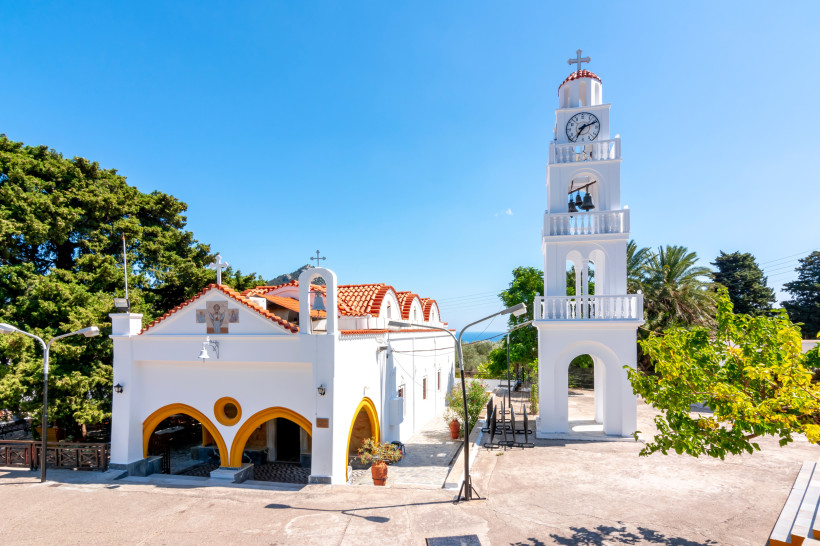 Weiße griechisch-orthodoxe Kirche auf Rhodos mit Glockenturm, rotem Ziegeldach und Palmen unter blauem Himmel