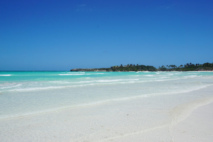 Panoramablick auf den traumhaften Strand auf Kuba mit türkisblauem Meer, sanften Wellen, weißem Sandstrand und Palmen im Hintergrund unter klarem, blauem Himmel.