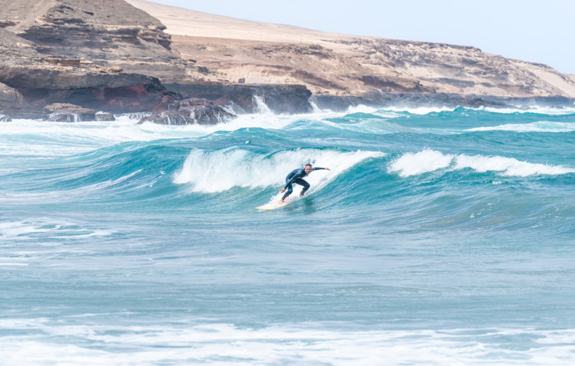 Surfer reitet eine Welle vor der rauen Felsküste von Fuerteventura bei strahlendem Sonnenschein