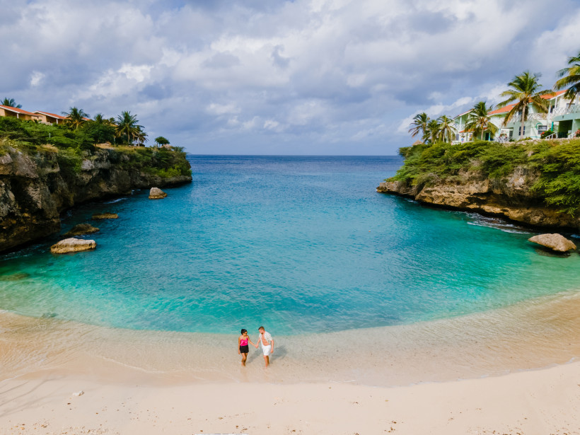 Curaçao - Playa Lagun Kleine, geschützte Bucht auf Curaçao mit klarem türkisblauem Wasser. Zwei Personen gehen am hellen Sandstrand entlang. Links und rechts säumen grüne Klippen mit Palmen und Häusern den Eingang zur Bucht, darüber ein teils bewölkter Himmel.