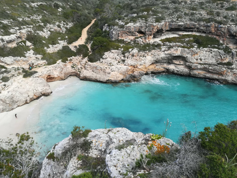 Mallorca - Cala Marmols Eine versteckte Bucht mit türkisfarbenem Wasser, umgeben von hohen, zerklüfteten Felswänden auf Mallorca. Eine kleine sandige Bucht liegt eingebettet zwischen den Felsen, der Strand ist fast menschenleer. Ein schmaler Pfad führt durch die grüne Vegetation