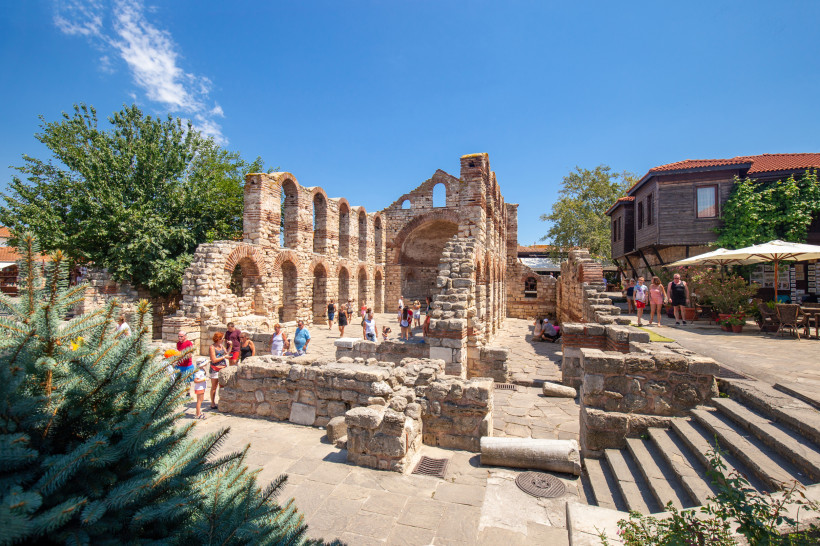 Ruinen der Kirche des Heiligen Sofia in Nessebar. Die romanisch-byzantinischen Mauern mit hohen Rundbögen bestehen aus Stein und Ziegel. Besucher schlendern zwischen den Säulen, im Hintergrund stehen Bäume und traditionelle Holzhäuser mit Restaurants.