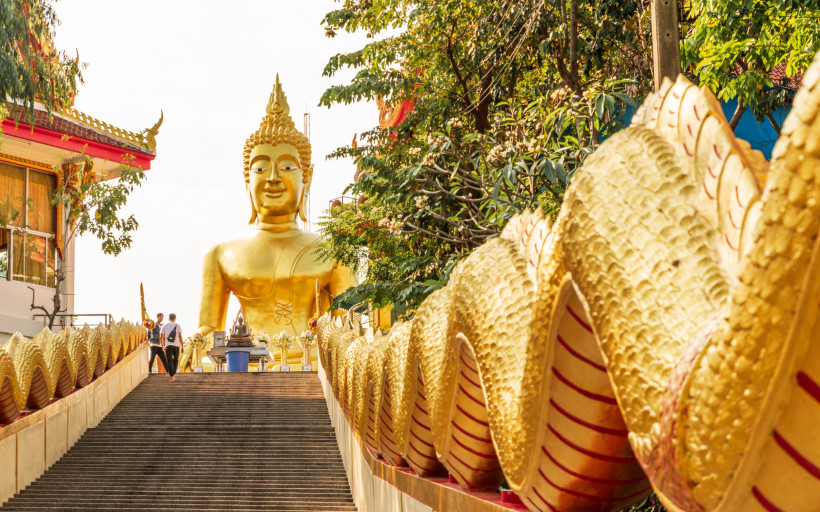 Goldene Buddha-Statue auf dem Tempelberg von Pattaya – Wat Phra Yai Goldene Buddha-Statue auf der Treppe von Wat Phra Yai in Pattaya, Thailand, flankiert von goldenen Schlangenverzierungen und Bäumen