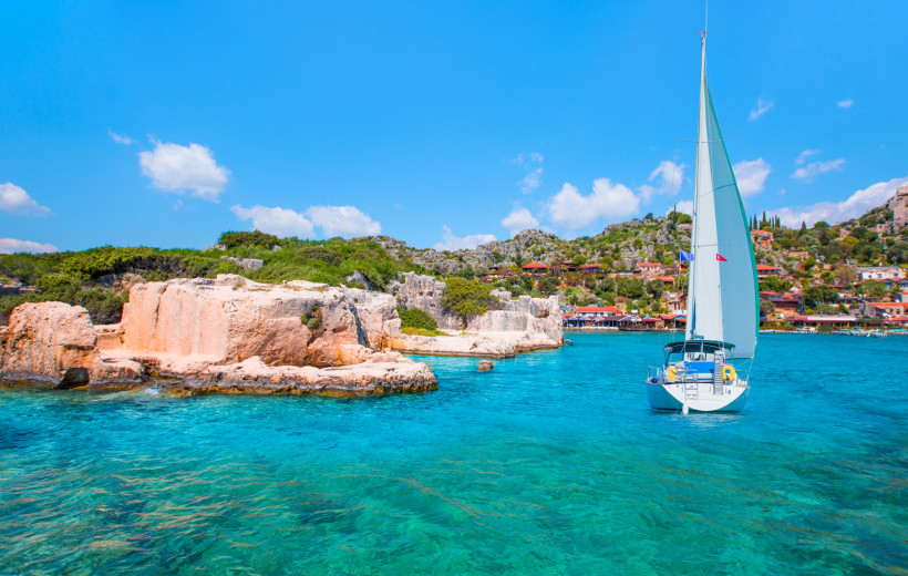 Ein weißes Segelboot fährt über türkisblaues Wasser entlang einer felsigen Küste mit grüner Vegetation. Im Hintergrund liegt ein malerisches Dorf mit traditionellen Häusern am Hang, unter strahlend blauem Himmel.