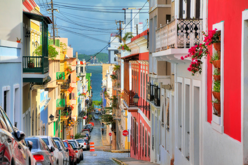 Bunte Häuserzeile mit Balkonen an einer steilen Straße in Old San Juan, Puerto Rico, mit Blick auf das Meer