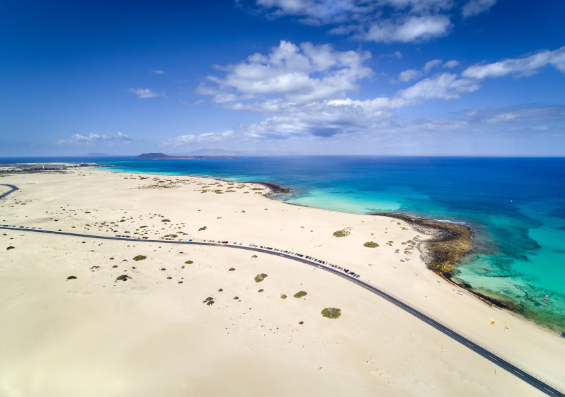 Luftaufnahme der Dünenlandschaft und Küste bei Corralejo mit Straße entlang des hellen Sandstrands und türkisfarbenem Meer