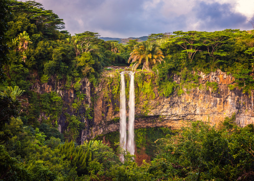 Dieses Bild zeigt einen spektakulären Wasserfall, der sich von einer hohen, felsigen Klippe in die Tiefe stürzt. Die Szenerie ist umgeben von dichter, tropischer Vegetation mit hohen Bäumen, Farnen und Palmen, die in leuchtendem Grün erstrahlen. Die Felsw