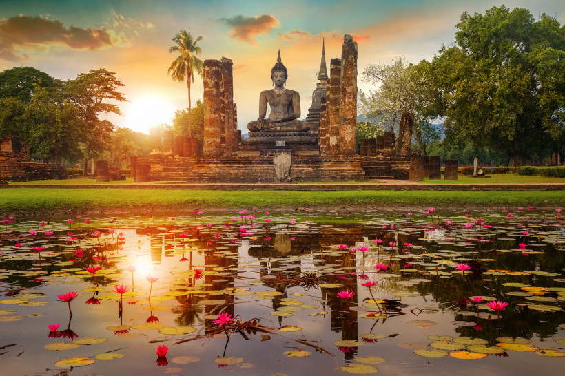 Sukhothai Historical Park bei Sonnenaufgang – Buddha-Statue Buddha-Statue im Sukhothai Historical Park in Thailand bei Sonnenaufgang, umgeben von einem Seerosenteich und historischen Tempelruinen.