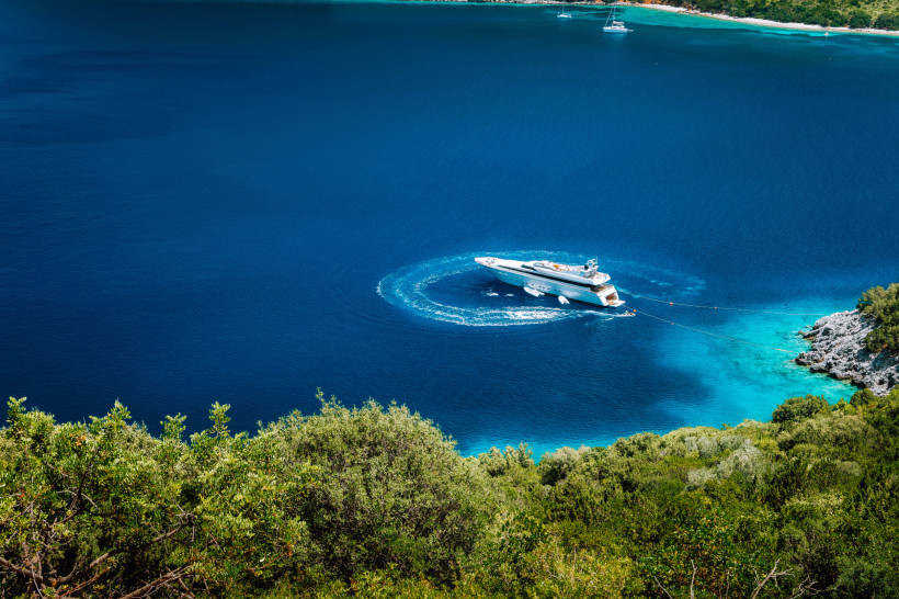 Exklusiver Moment auf dem Wasser: Eine elegante Luxusyacht fährt durch eine smaragdgrüne Bucht, das türkisfarbene Wasser wirbelt in sanften Kreisen um den Rumpf. Die Szene wird eingerahmt von grüner Vegetation und felsiger Küste – mediterraner Luxus pur.
