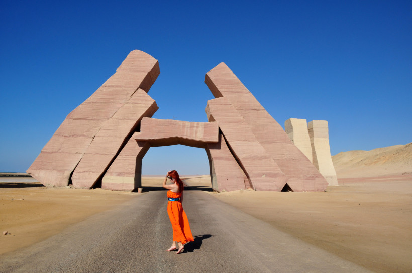 Ägypten - Sharm el Sheikh Eine Frau mit langen roten Haaren und einem leuchtend orangefarbenen Kleid steht barfuß auf einer Straße mitten in der Wüste. Im Hintergrund erhebt sich ein monumentales, modernes Steintor aus mehreren schräg stehenden, kantigen Blöcken in rötlichen Farbt