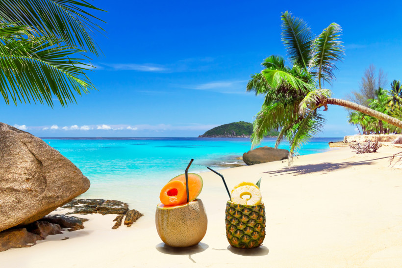 Zwei tropische Cocktails in einer Melone und einer Ananas am weißen Sandstrand mit Palmen und türkisblauem Meer