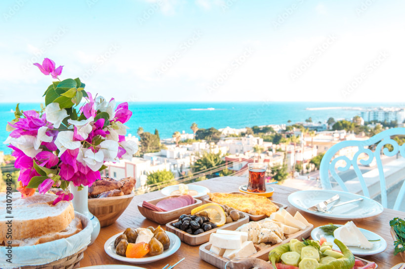 Traditionelles Frühstück auf einer Terrasse mit Meerblick in der Türkei, gedeckter Tisch mit regionalen Speisen, Blumen und Blick auf Küstenort und Mittelmeer