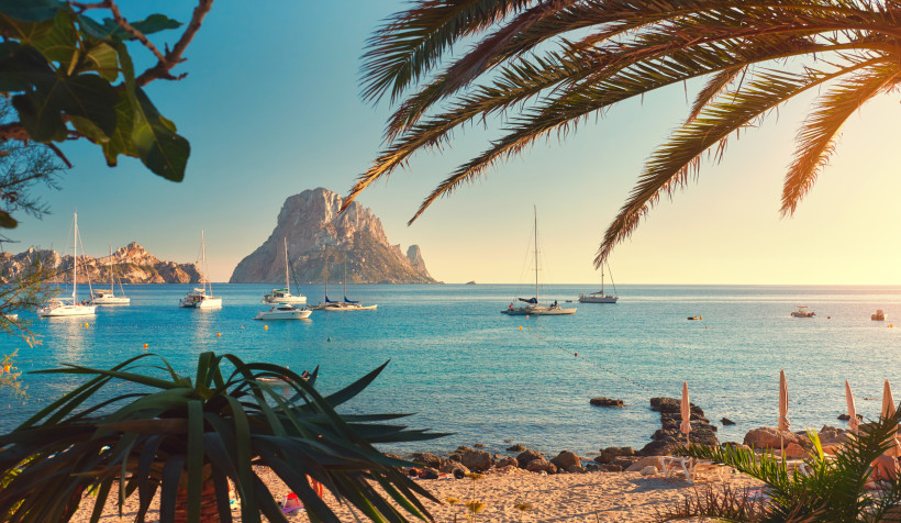 Es Vedra vor Ibiza bei ruhiger See mit vor Anker liegenden Segelbooten Blick auf die Felseninsel Es Vedra vor Ibiza mit Segelbooten auf dem Meer und Palmen im Vordergrund