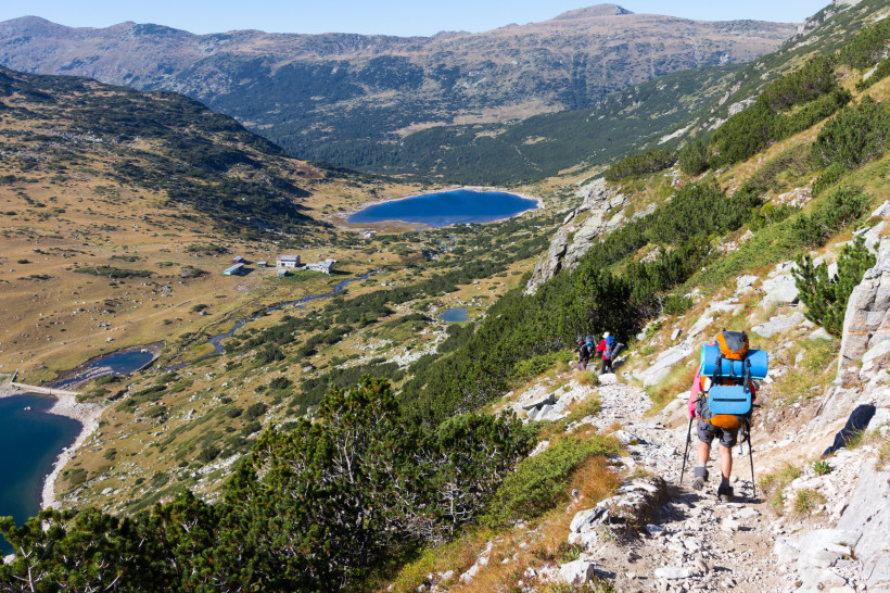 Bulgarien Wanderer mit Rucksack und Trekkingstöcken auf einem steinigen Bergpfad. Der Weg führt durch das grüne Rila-Gebirge in Bulgarien. Im Hintergrund liegen mehrere klare, blaue Bergseen eingebettet in eine weite, offene Landschaft mit sanften Hügeln und verstr