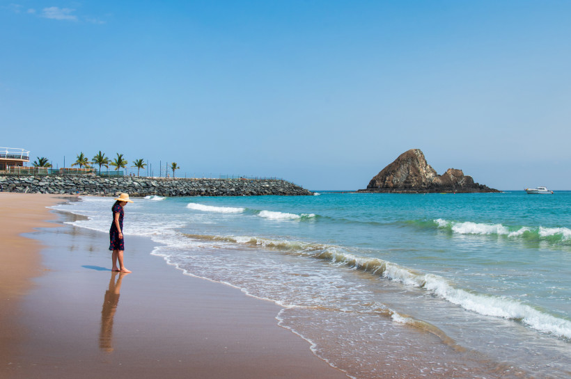 Fujairah, Strandferien Frau mit Sonnenhut steht am Sandstrand und schaut auf das Meer mit einer Felseninsel im Hintergrund